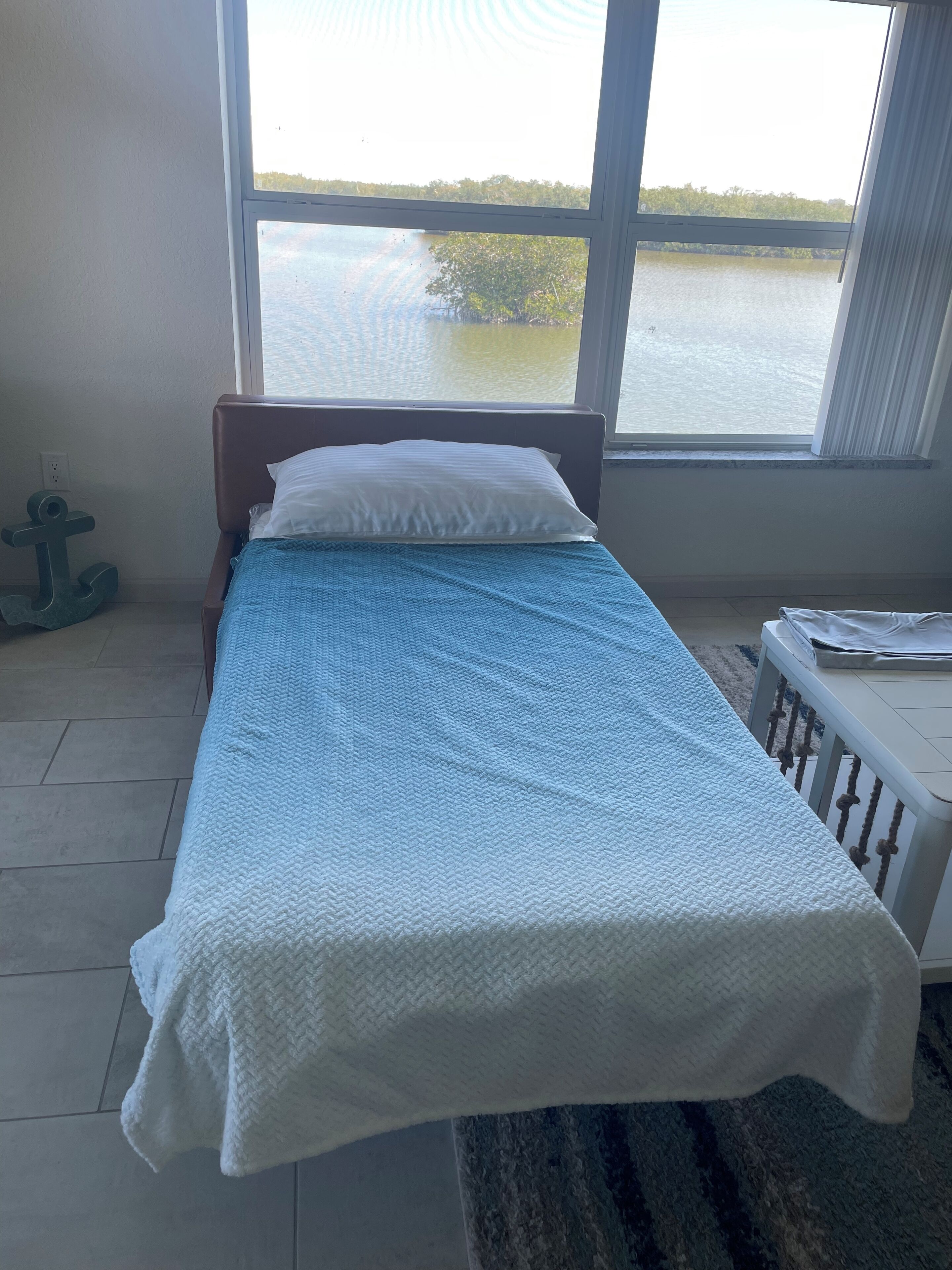 Een bed met een blauw-witte deken, een wit kussen en uitzicht op een waterlichaam door een raam.
