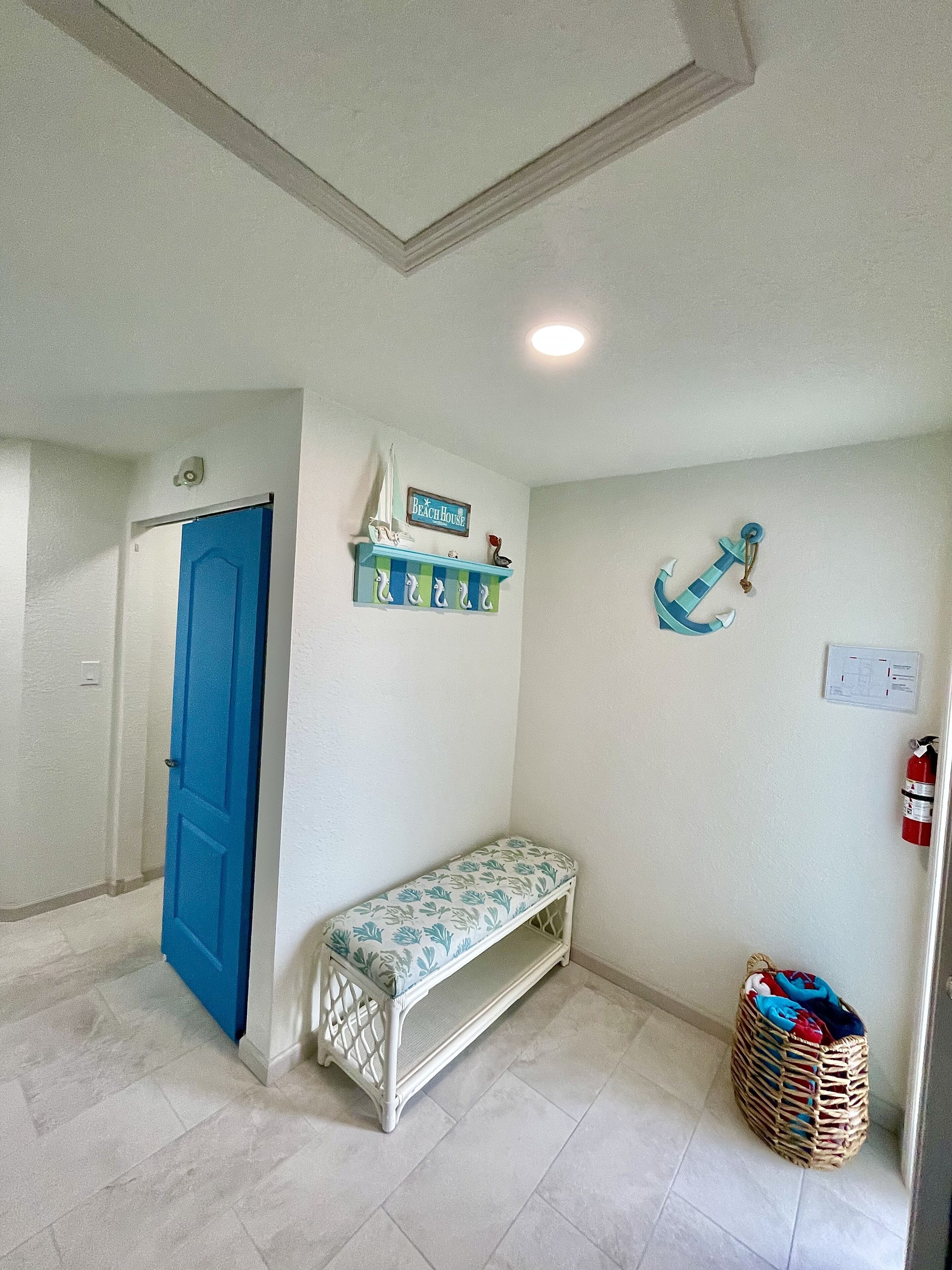Un pasillo con una puerta azul, un banco, una estantería con adornos náuticos y un extintor.