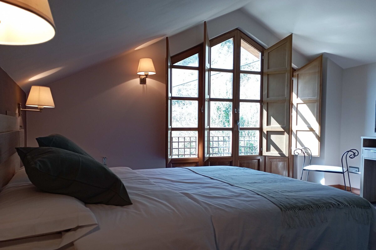 A bedroom with a bed, two wall-mounted lamps, a window with a view of trees, and a desk with a chair.