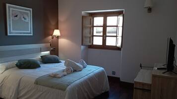 A neatly made bed with pillows and rolled towels, a bedside lamp, a window with wooden shutters, and a television on a wooden stand.