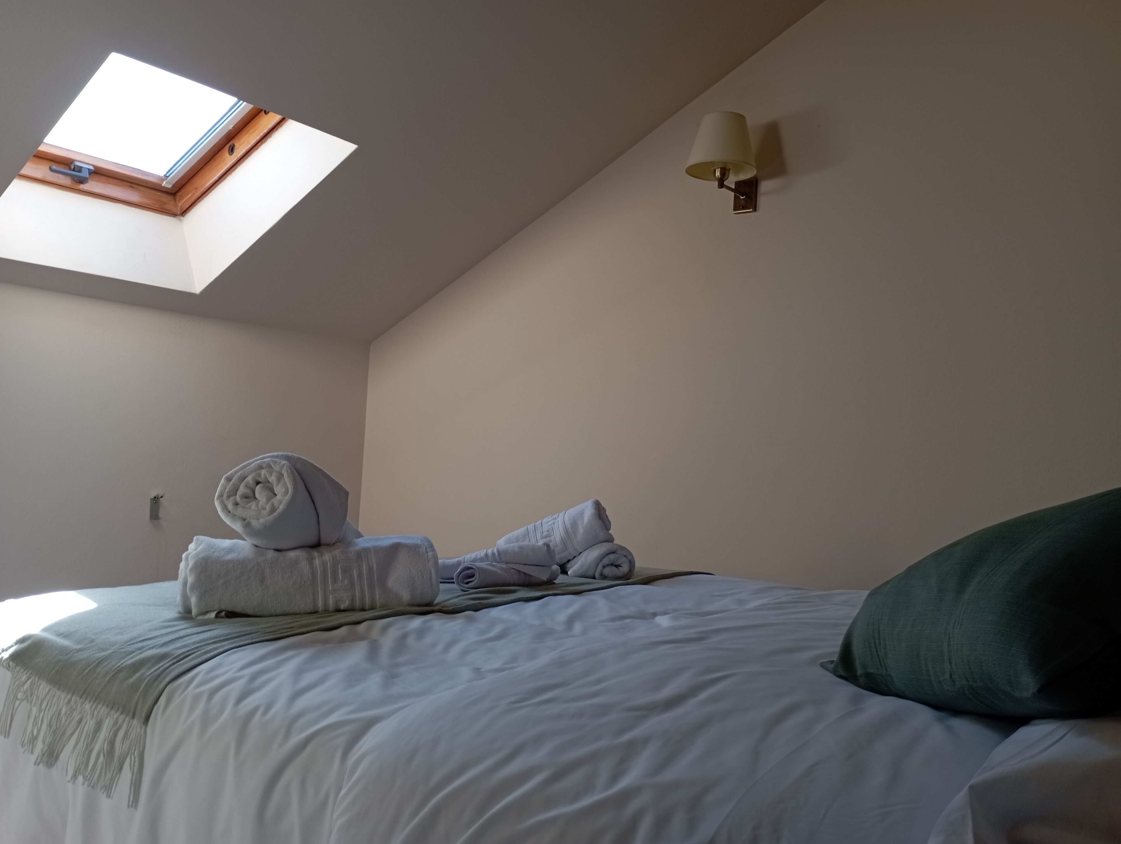 A bedroom with a skylight, a bed with white bedding, a rolled towel, and a wall-mounted lamp.