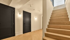 Una escalera de madera con peldaños que conduce a un rellano con dos puertas negras y luces fijadas a la pared.