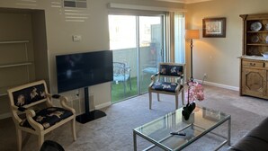 Een woonkamer met een flatscreen-tv, een glazen salontafel, een bank en een stoel met een decoratief kussen. Er is een schuifdeur naar een patio met buitenmeubilair.