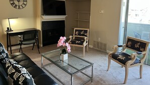 Een woonkamer met een bank, een glazen salontafel, een televisie, een open haard en twee fauteuils met gestreepte kussens.