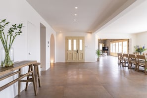 Een ruime kamer met een houten tafel en stoelen, een groot raam en een deur naar een ander vertrek.