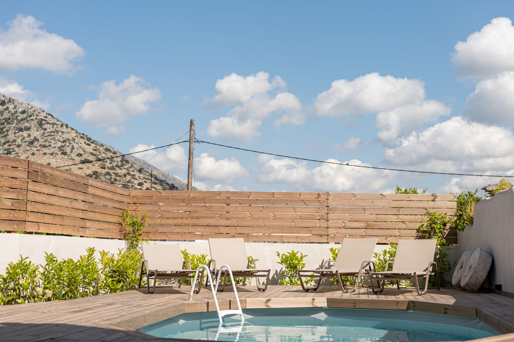 Zona de piscina con terraza de madera, sillones de descanso y montañas al fondo.