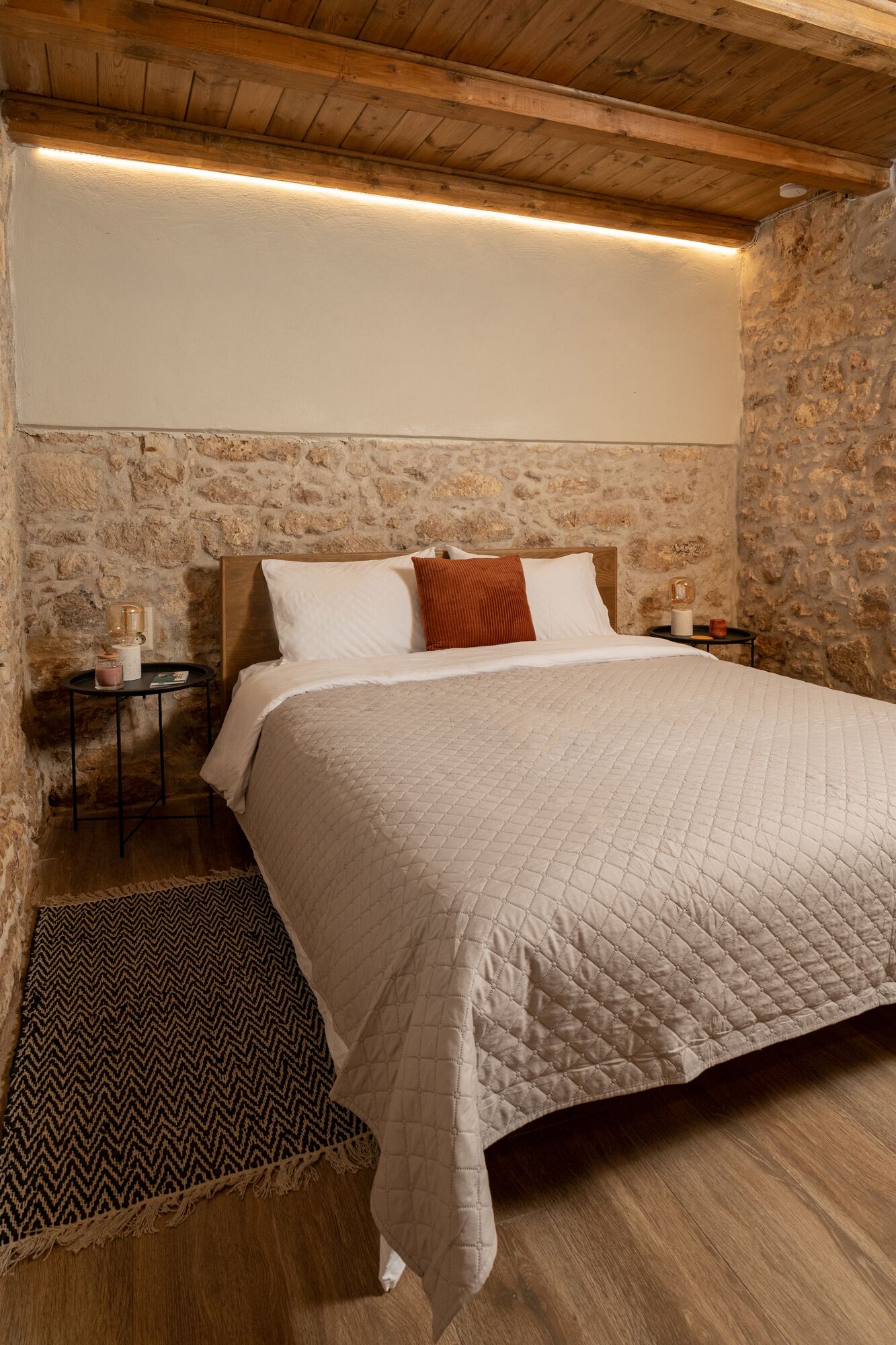 A bedroom with a large bed, stone walls, wooden beams, a small bedside table with a lamp, and a rug.
