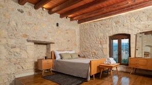 Une chambre avec un mur en pierre, des poutres en bois au plafond, un lit avec une tête de lit en bois, une table de chevet, une commode et une vue à travers une porte.