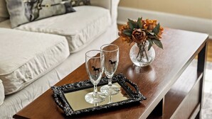 A wooden table with a mirror tray holding two wine glasses, a vase with dried flowers, and a sofa with a floral pillow in the background.