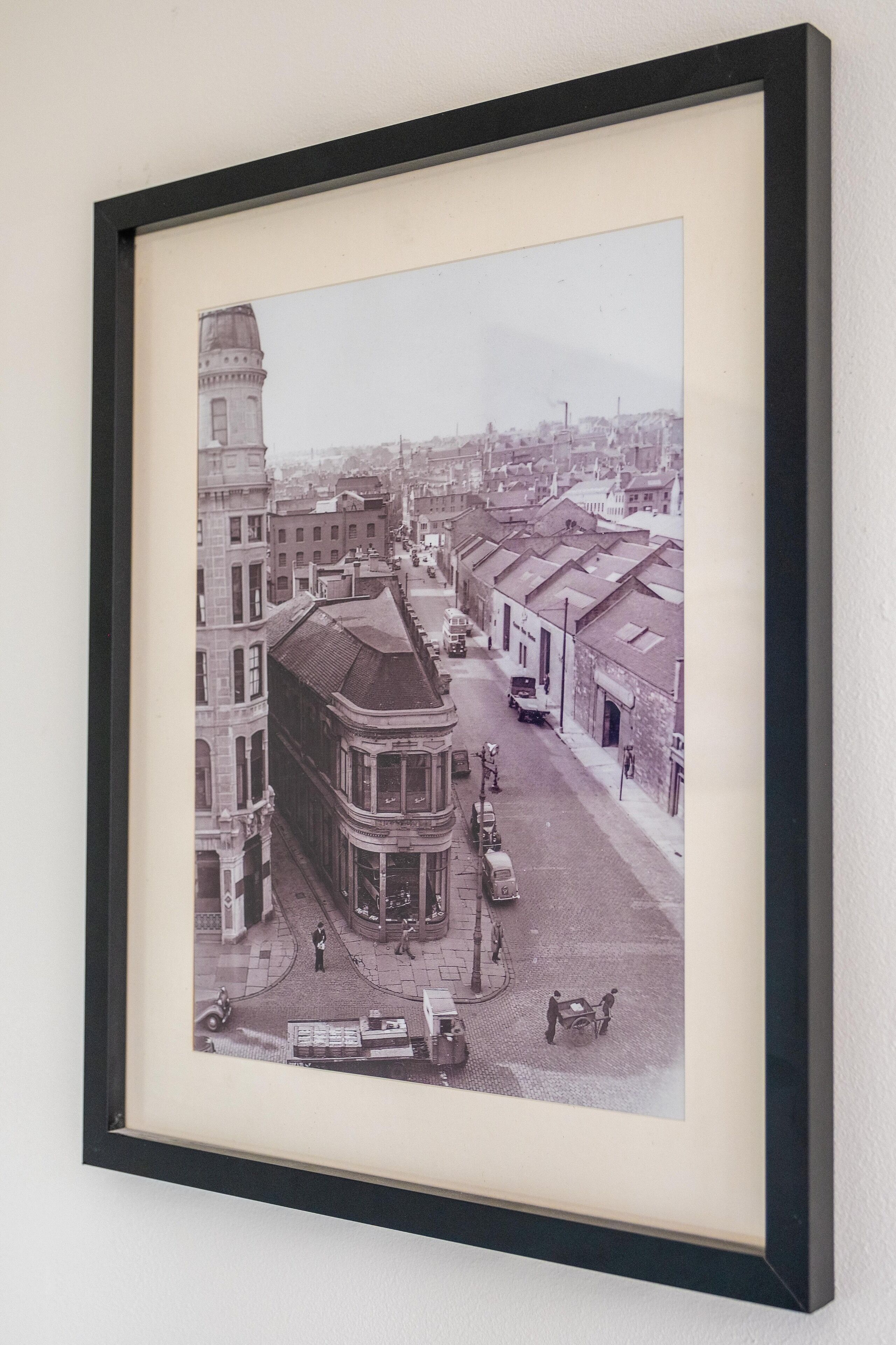 Fotografía histórica enmarco que muestra una escena de calle con edificios, una torre destacada y vehículos del siglo XX.