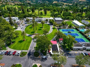 Vista aérea de un campo de golf con cancha de tenis, zona de estacionamiento y follaje circundante.