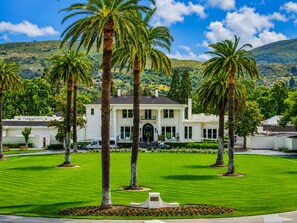 A white two-story house with a symmetrical front, surrounded by a well-maintained lawn and palm trees, set against a hilly backdrop. - Relaxing wine country condo at Silverado in Napa (Napa)