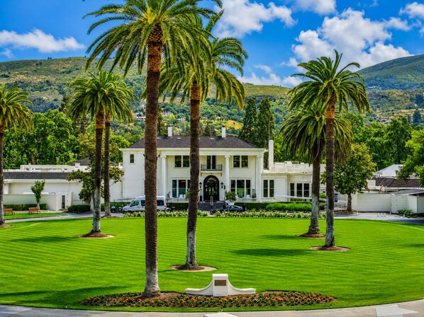 A white two-story house with a symmetrical front, surrounded by a well-maintained lawn and palm trees, set against a hilly backdrop.