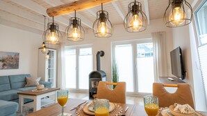 A dining area with a wooden table, chairs, and a glass of orange juice. There is a sofa and a fireplace in the background. - Ferienhaus Marschengrün Büsum (Büsum)