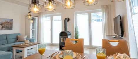 A dining area with a wooden table, chairs, and a glass of orange juice. There is a sofa and a fireplace in the background.