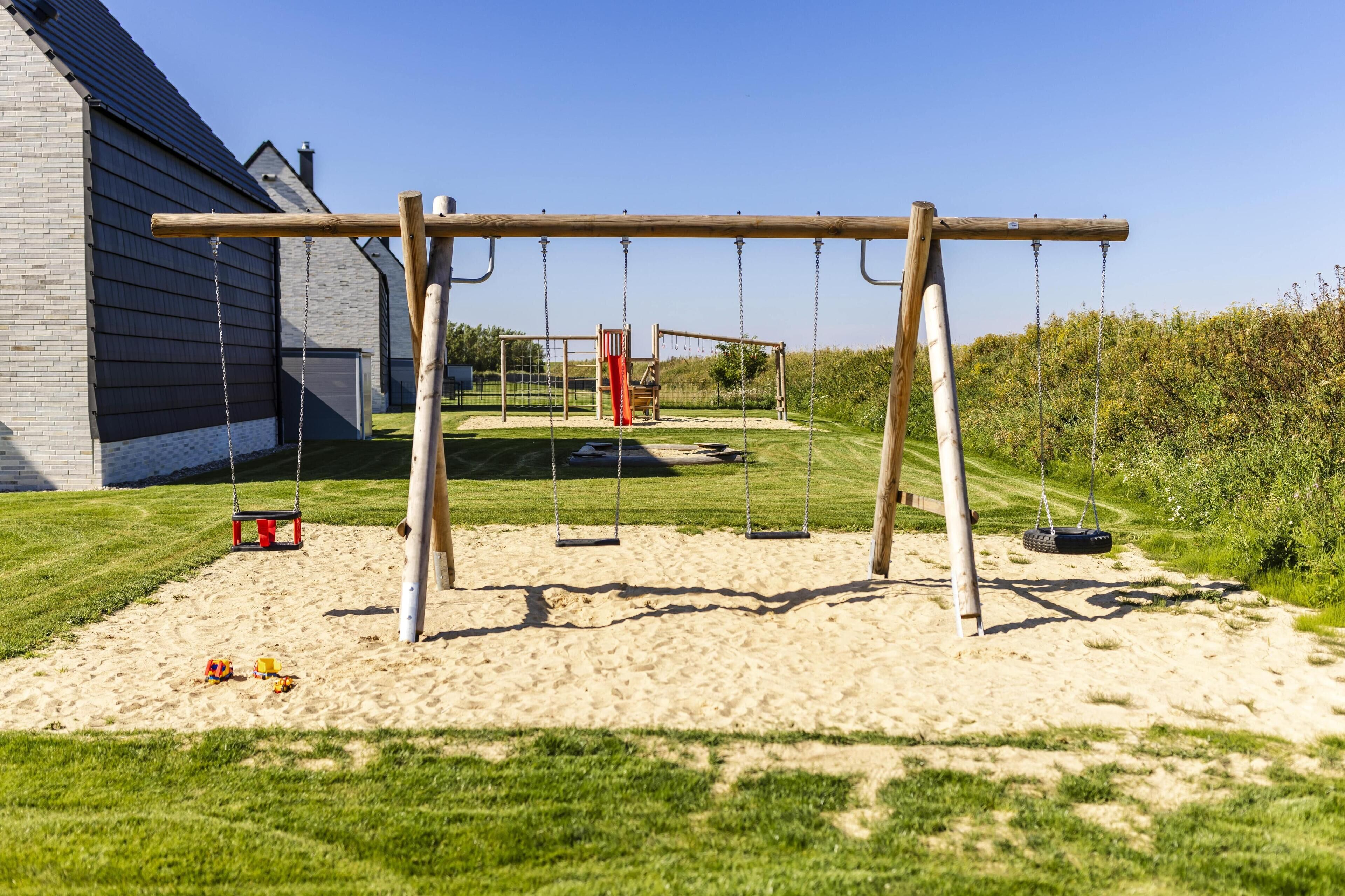 Ein Spielplatz mit Schaukeln und einer Fahrradschaukel, vor einer Gebäudefassade und einer grünen Wiese.