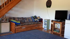 Un accogliente soggiorno con una scala in legno, un divano con cuscini decorati, una bacheca da freccette, un televisore e una libreria.