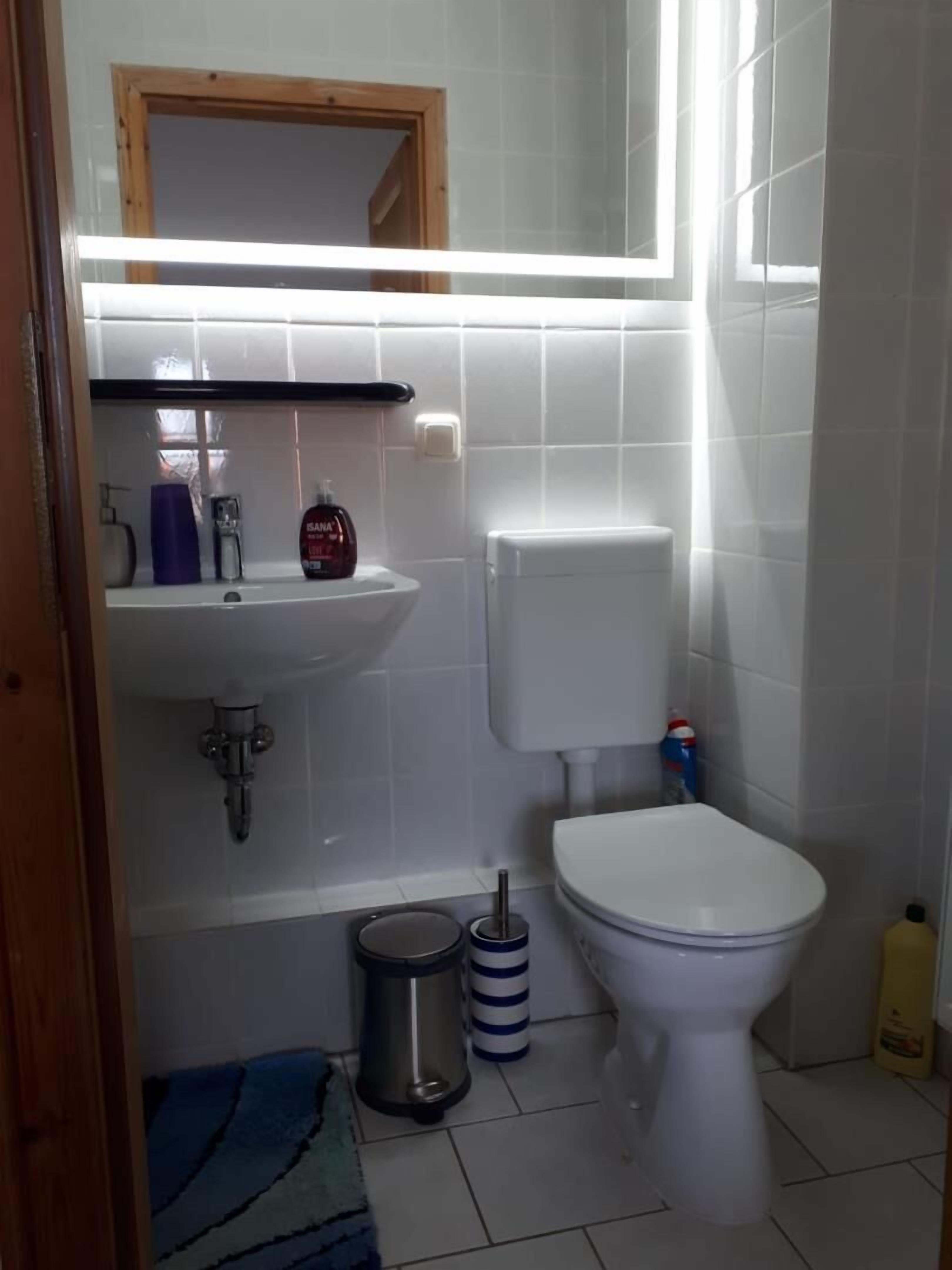 Un baño con inodoro, lavamanos y espejo sobre él. Hay un cubo de basura y una botella de producto de limpieza en el suelo.