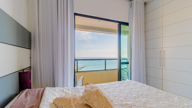 A bedroom with a large bed, a view of the sea through a sliding door, and a balcony with a chair. - RV09 Apto Vista Mar - Rio Vermelho (Salvador)