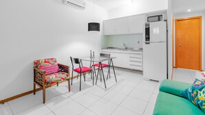 A compact kitchen with white cabinets, a microwave, and a refrigerator. There is a dining table with chairs, a sofa, and a chair with a colorful cushion. - RV09 Apto Vista Mar - Rio Vermelho (Salvador)