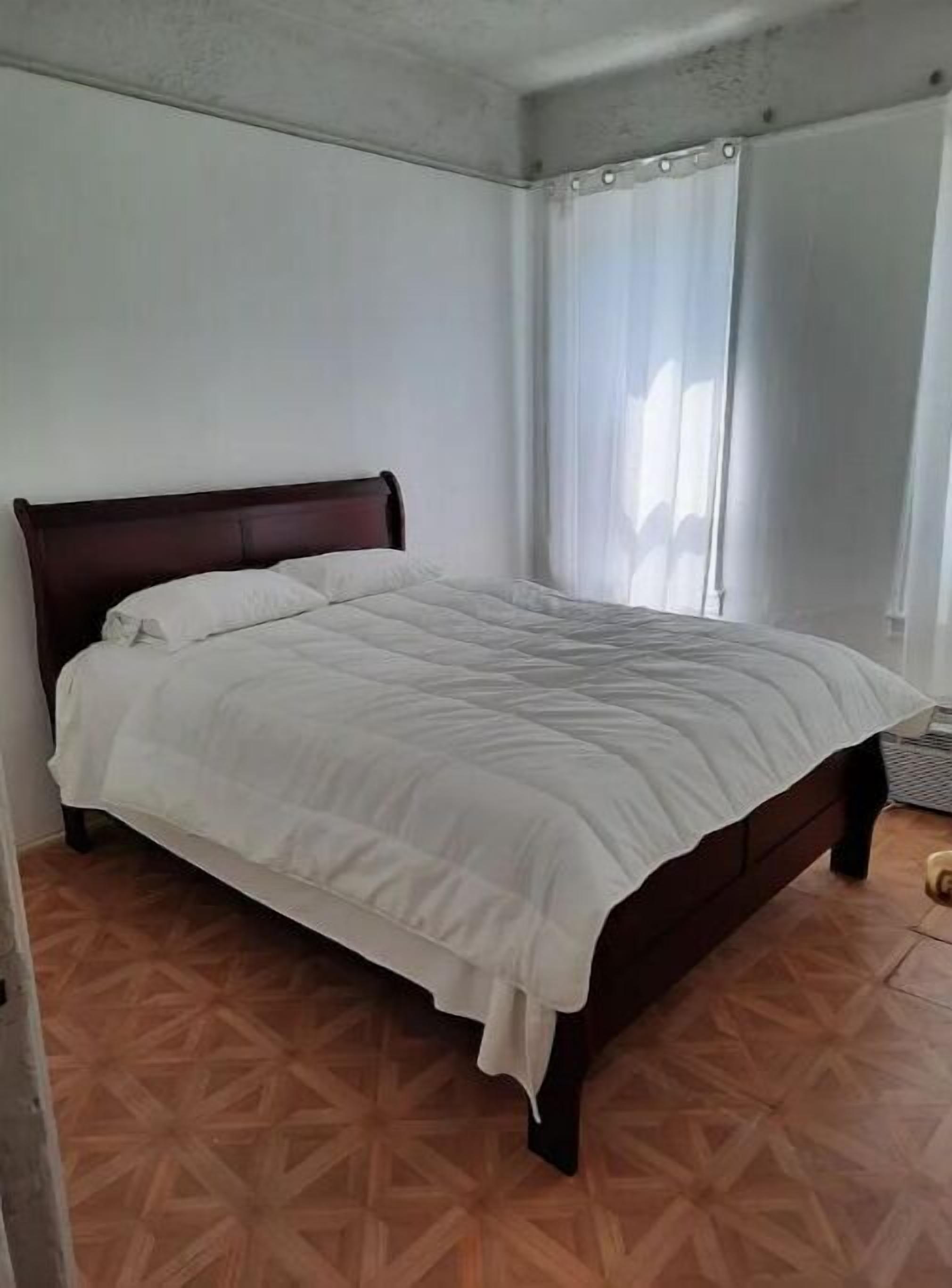 Un lit simple dans une pièce dont le sol est recouvert d’un motif décoratif, les murs sont blancs et il y a une fenêtre avec des rideaux.