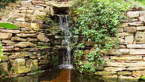 Une cascade de pierres se déversant dans un bassin aux eaux calmes, entourée de verdure et d’un mur de pierre.