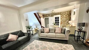 A living room with a grey sofa, a patterned rug, and a staircase leading to a dining area with a stone wall. - A Superb Size Cottage By The Canal (Torfaen)
