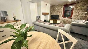 A modern living room with a stone accent wall, a grey sofa set, a wooden dining table, and a potted plant. - A Superb Size Cottage By The Canal (Torfaen)