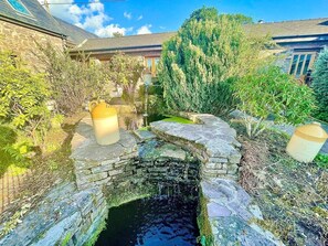 A stone bridge over a small water channel, surrounded by greenery and a building with a sloped roof.