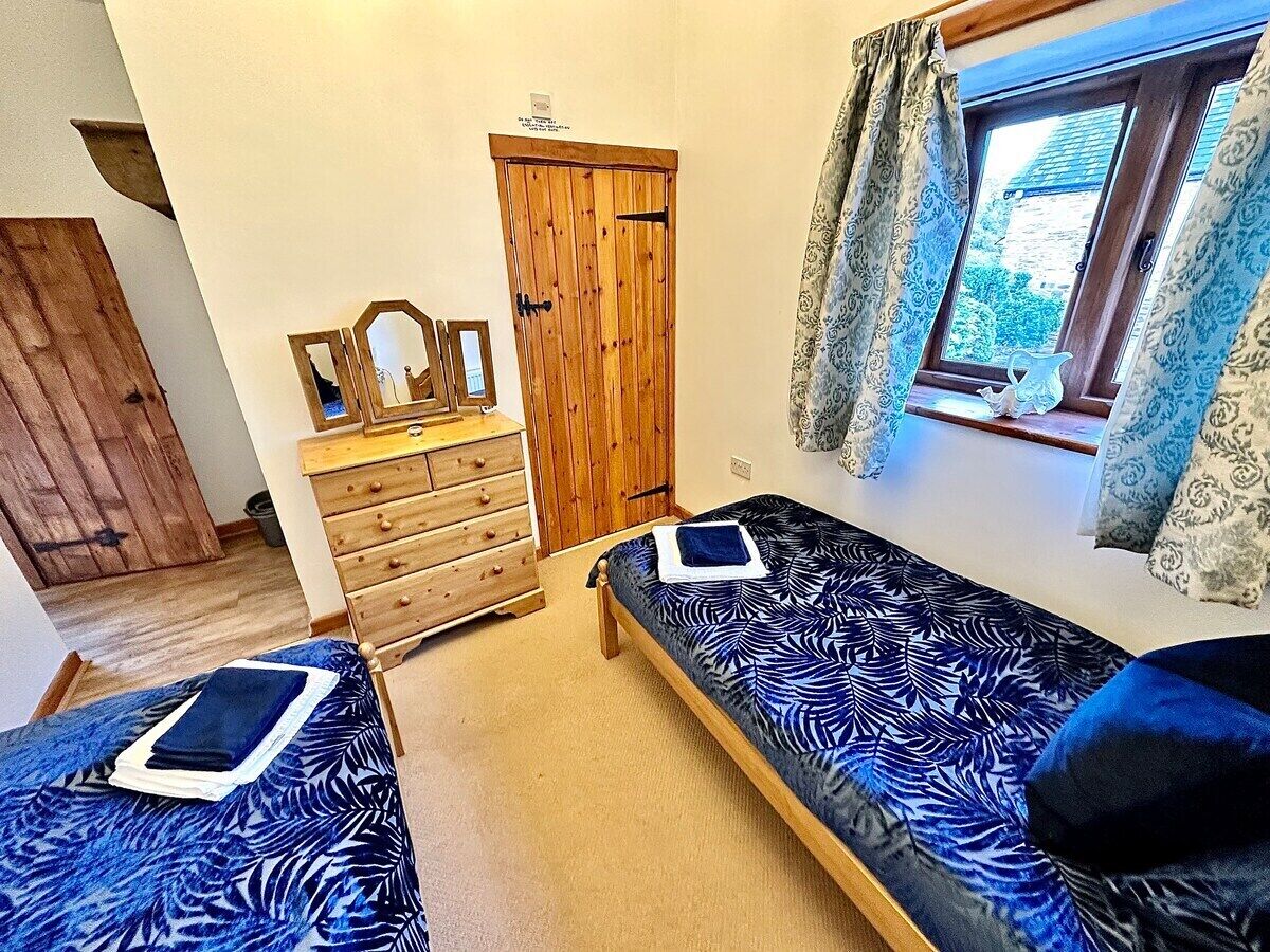 A room with two single beds, a wooden dresser, a door, and a window with curtains.