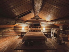 A log cabin bedroom with a bed, two bedside lamps, a washing machine, and a rustic wooden ceiling. - Cozy, Modern Log Cabin Getaway ~ 20 Min to Winter Park (Tabernash)