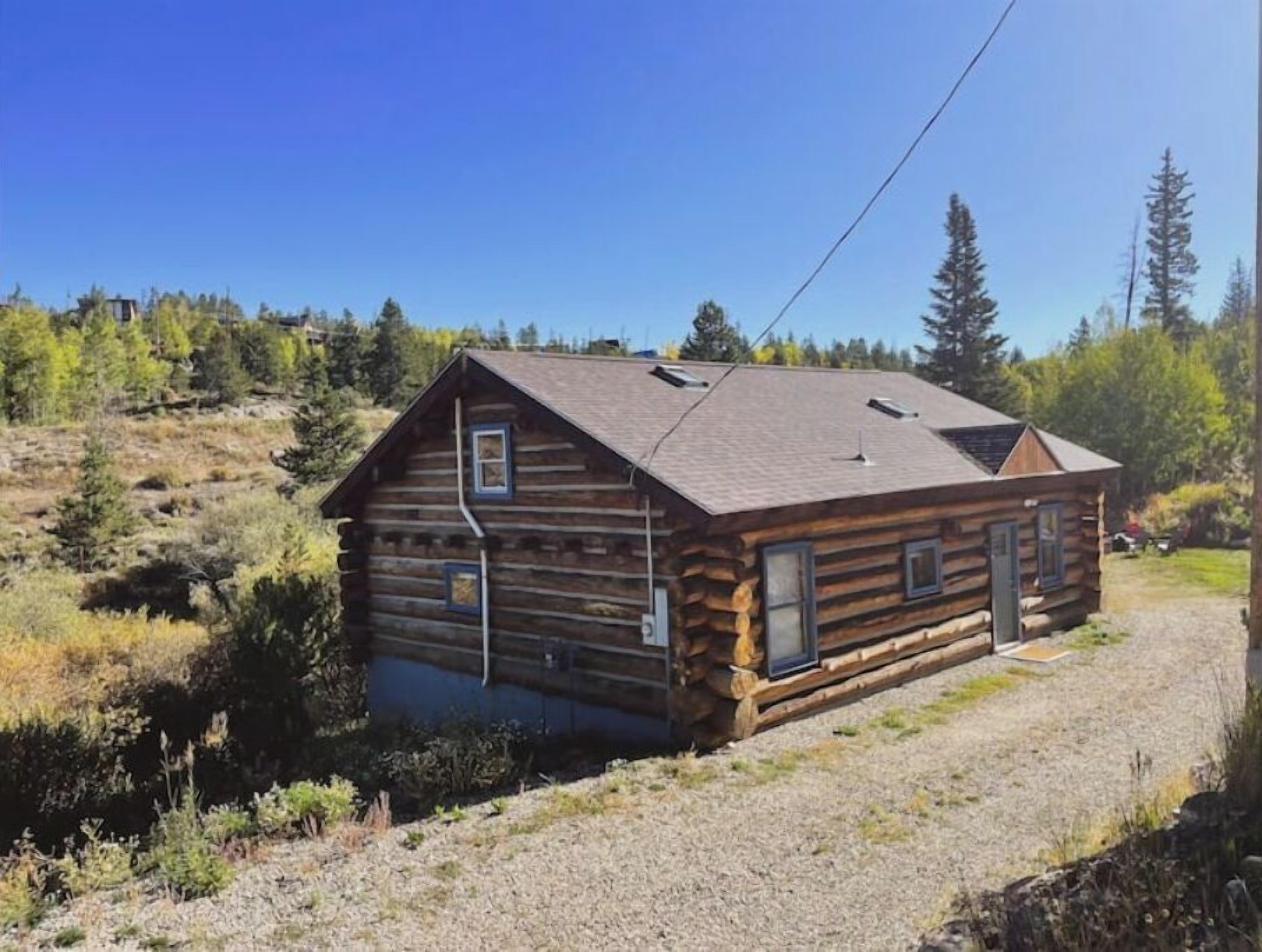 Uma cabana de tronco com telhado inclinado, um pequeno alpendre e uma entrada com calçamento de brita.