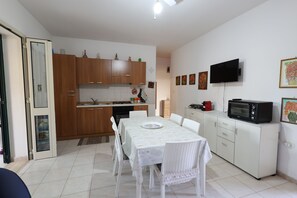 Uma cozinha compacta com armários de madeira, uma mesa de jantar com cadeiras brancas, micro-ondas e uma televisão na parede.
