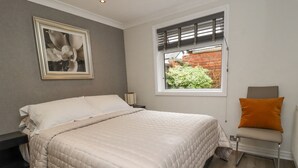 Un dormitorio con cama, mesita de noche, cortinas en la ventana, una silla e imagen en la pared.