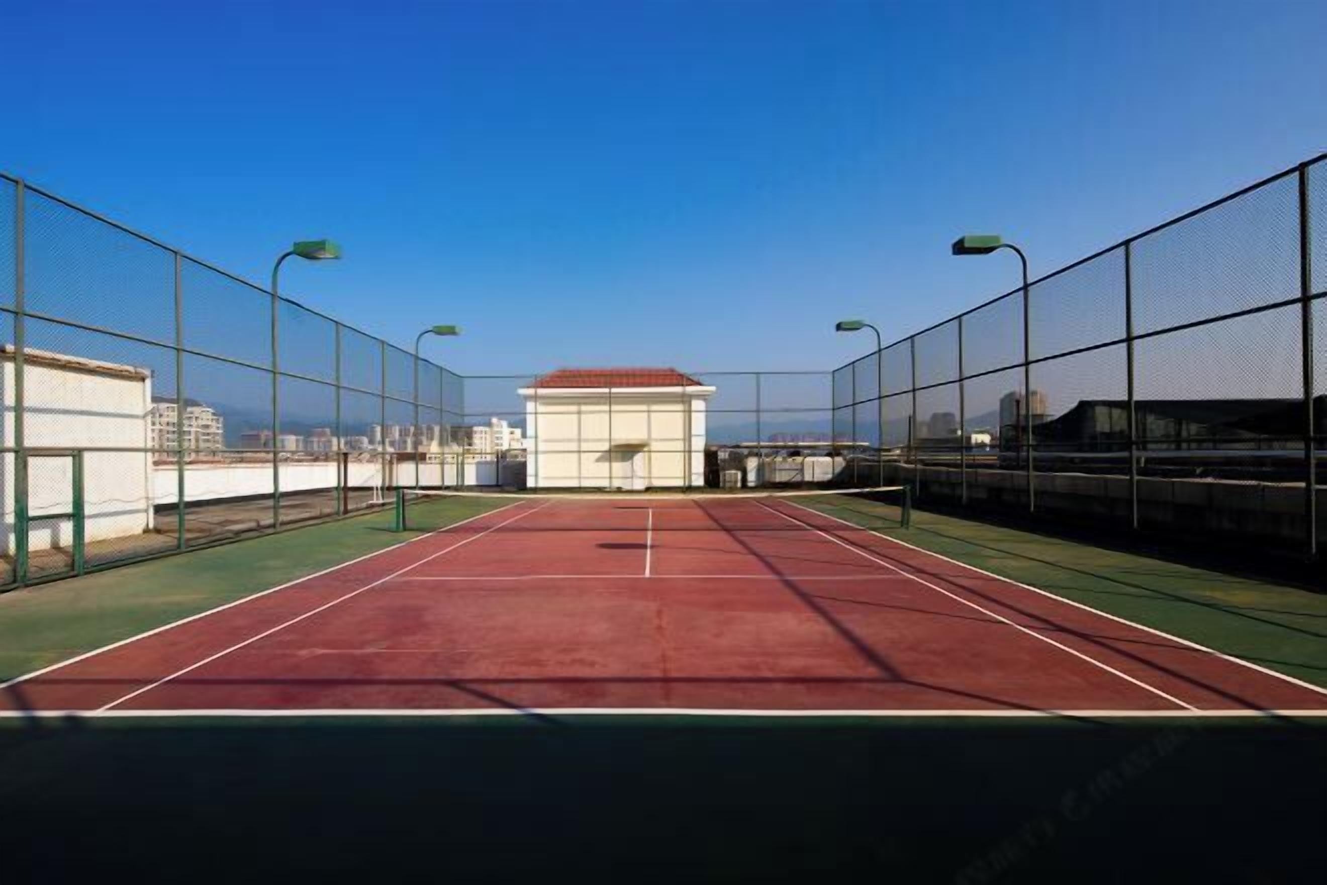 A tennis court with a net, surrounded by a fence, and a building with a red roof in the background.