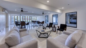 Un amplio espacio de sala con un conjunto de sofás, una mesa de centro y sillas. Hay ventiladores de techo, ventanas grandes y vistas al exterior.