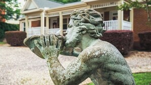 A statue of a muscular figure with clenched fists, set against a backdrop of a house with a porch and a well-maintained lawn.