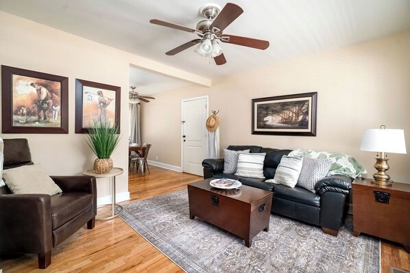 A living room with a dark brown leather sofa, a coffee table, and a ceiling fan. There are framed pictures on the walls and a plant on a side table.
