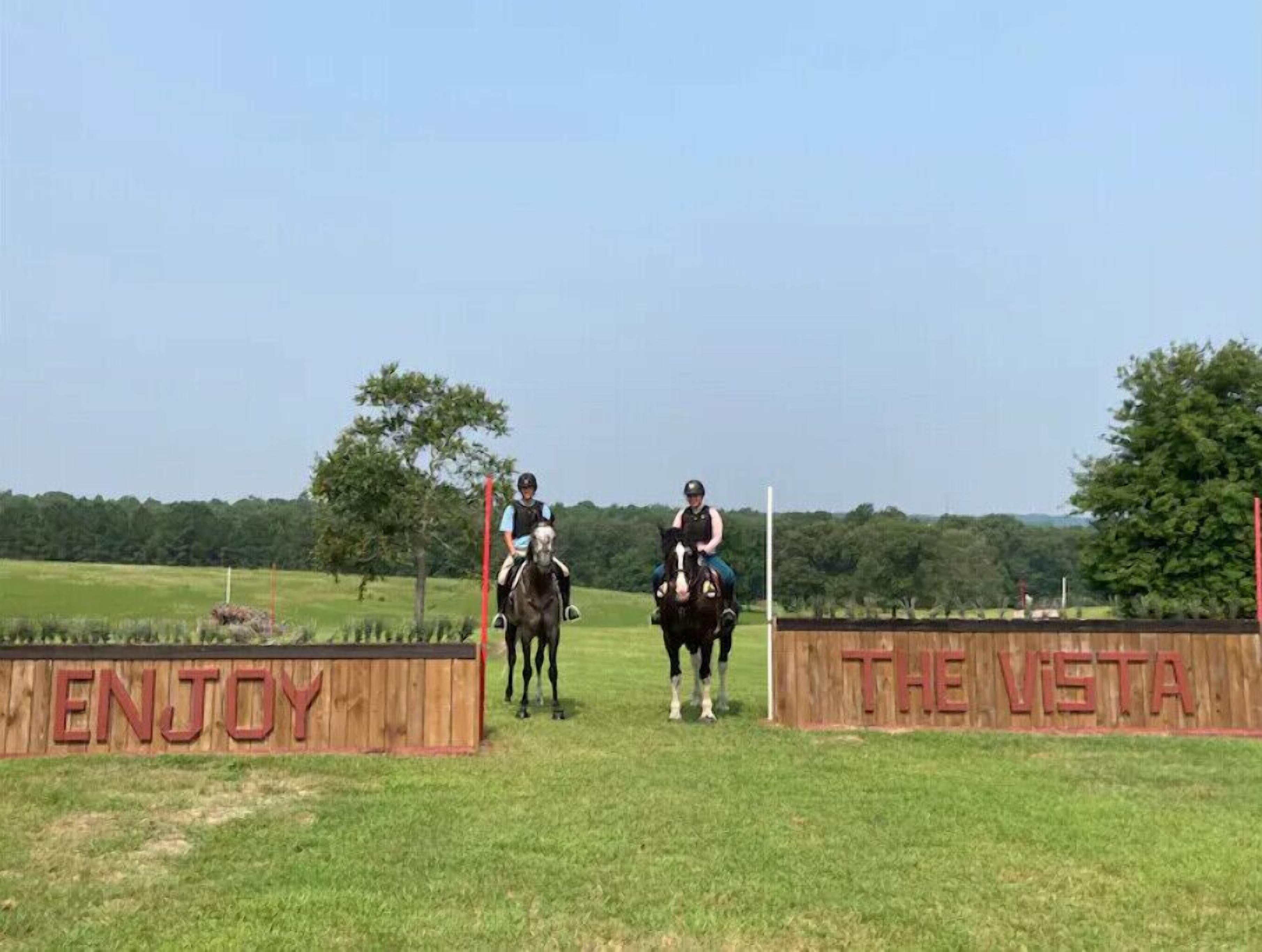 Two people on horseback at an outdoor equestrian course with wooden barriers and a sign that reads "ENJOY THE VISTA".