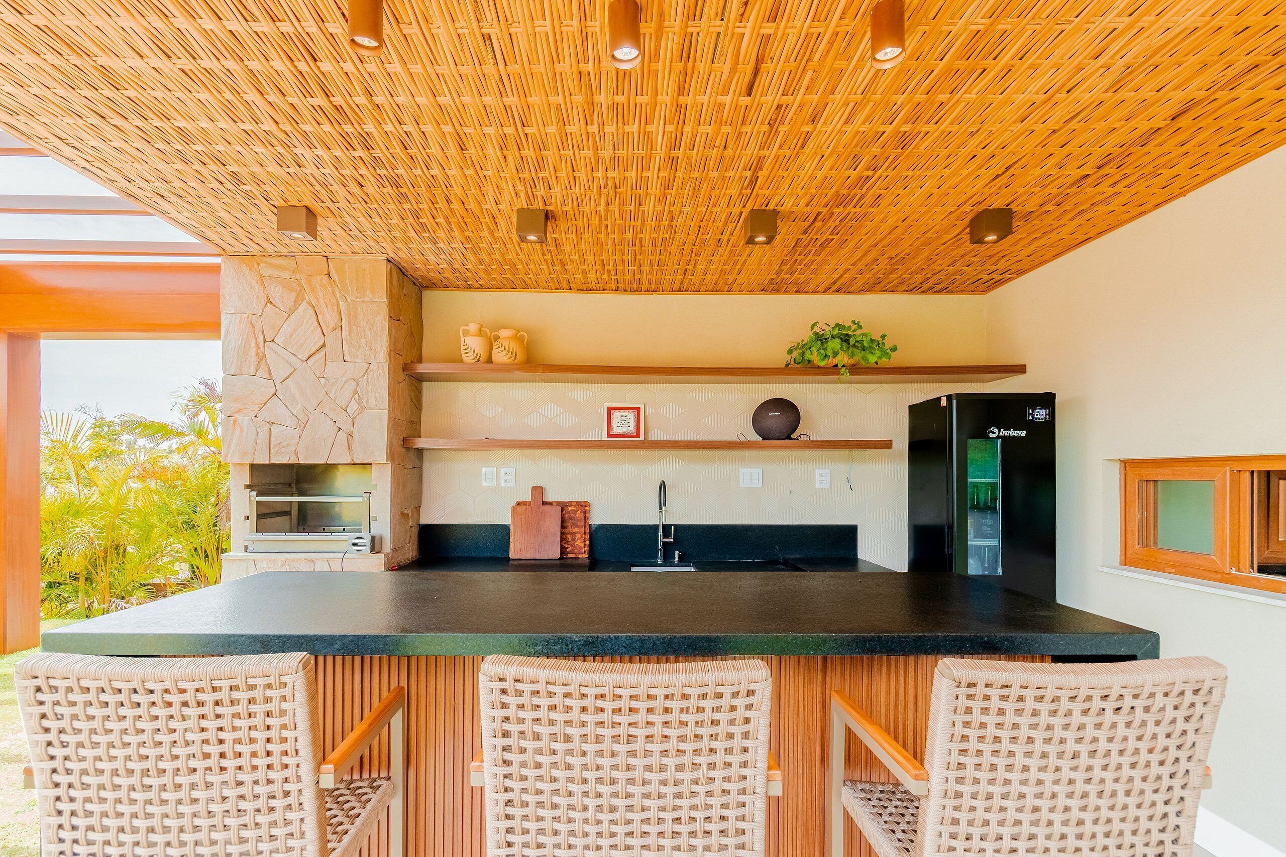 Una cocina moderna con techo de bambú, refrigerador negro y una zona de bar con sillas de mimbre.
