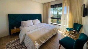 Une chambre d’hôtel avec un grand lit, une table de chevet, une chaise, une petite table avec une lampe et une fenêtre avec des rideaux.