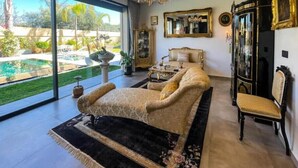 Un vaste salon doté d’une grande porte vitrée donnant sur une piscine, d’un canapé classique, d’un lustre et d’une commode ancienne.