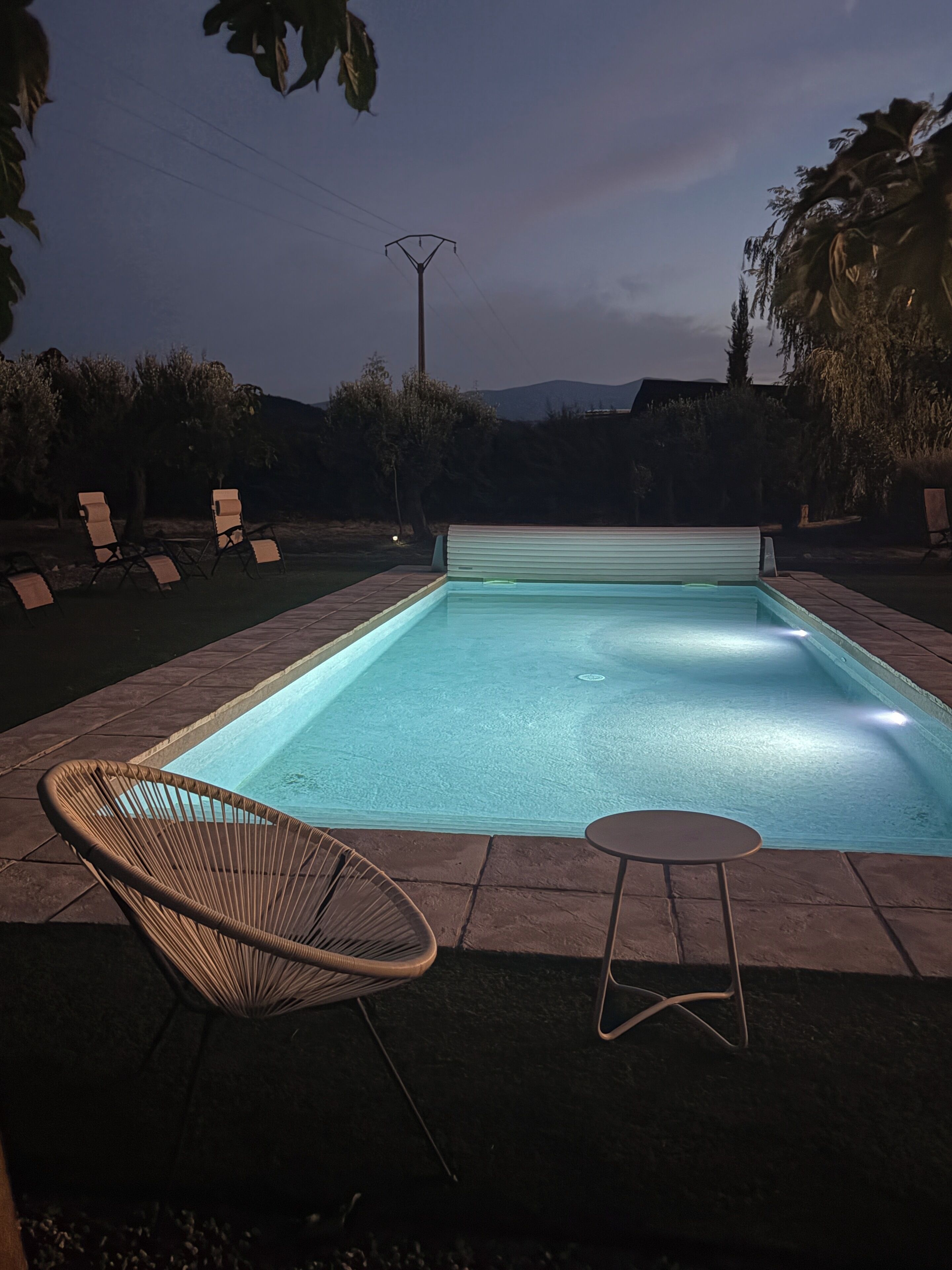 A rectangular swimming pool with a tiled edge, surrounded by a patio area with lounge chairs and a small round table.
