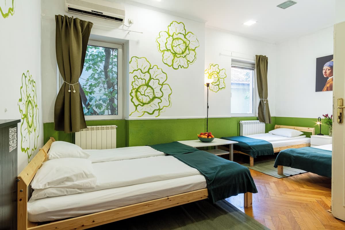 A hotel room with two single beds, a small table with a fruit bowl, a radiator, and green wall art.