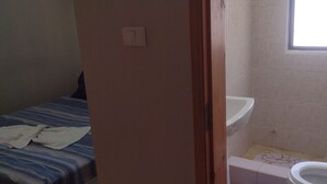 Ein kleiner, schwach beleuchteter Raum mit einem Bett, einem Lichtschalter, einem Fenster und einer Toilette.