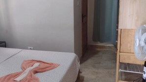 Ein Schlafzimmer mit Bett, Nachttisch, Ventilator und einer Tür zu einem anderen Raum.