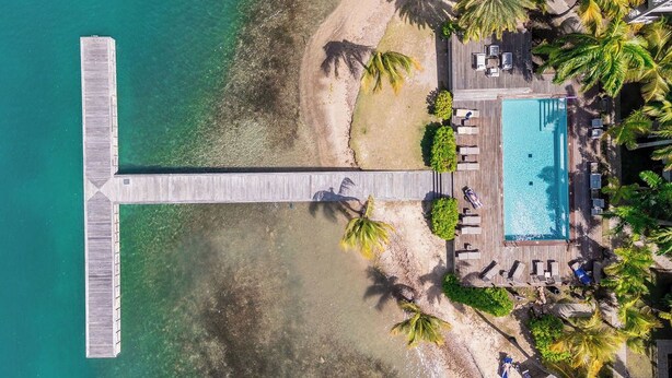 Vue aérienne d’un complexe hôtelier doté d’une piscine, de chaises longues, de palmiers et d’une jetée en bois.