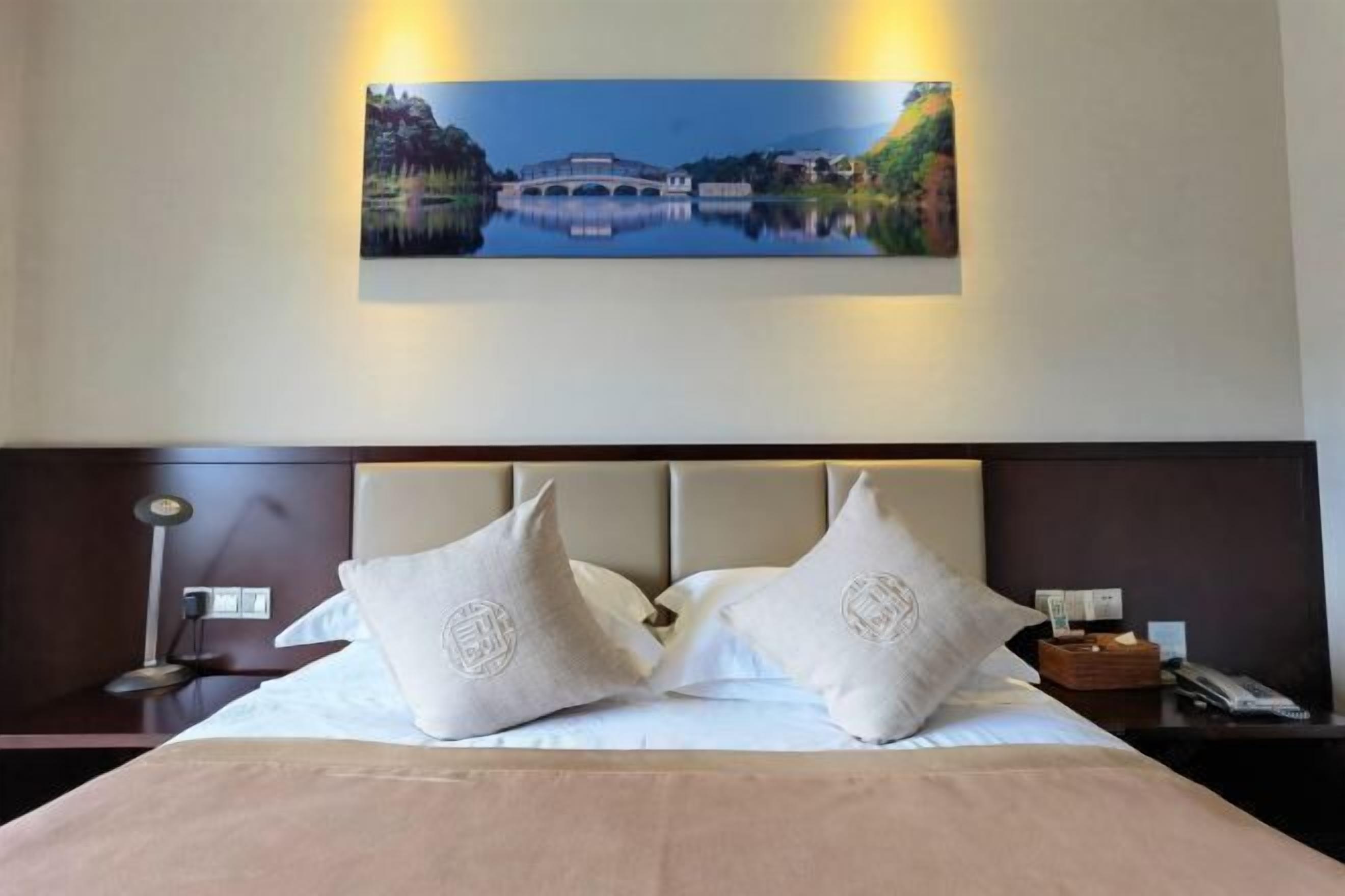 A hotel room with a bed, two pillows, a nightstand, a lamp, and a painting of a bridge over a lake.
