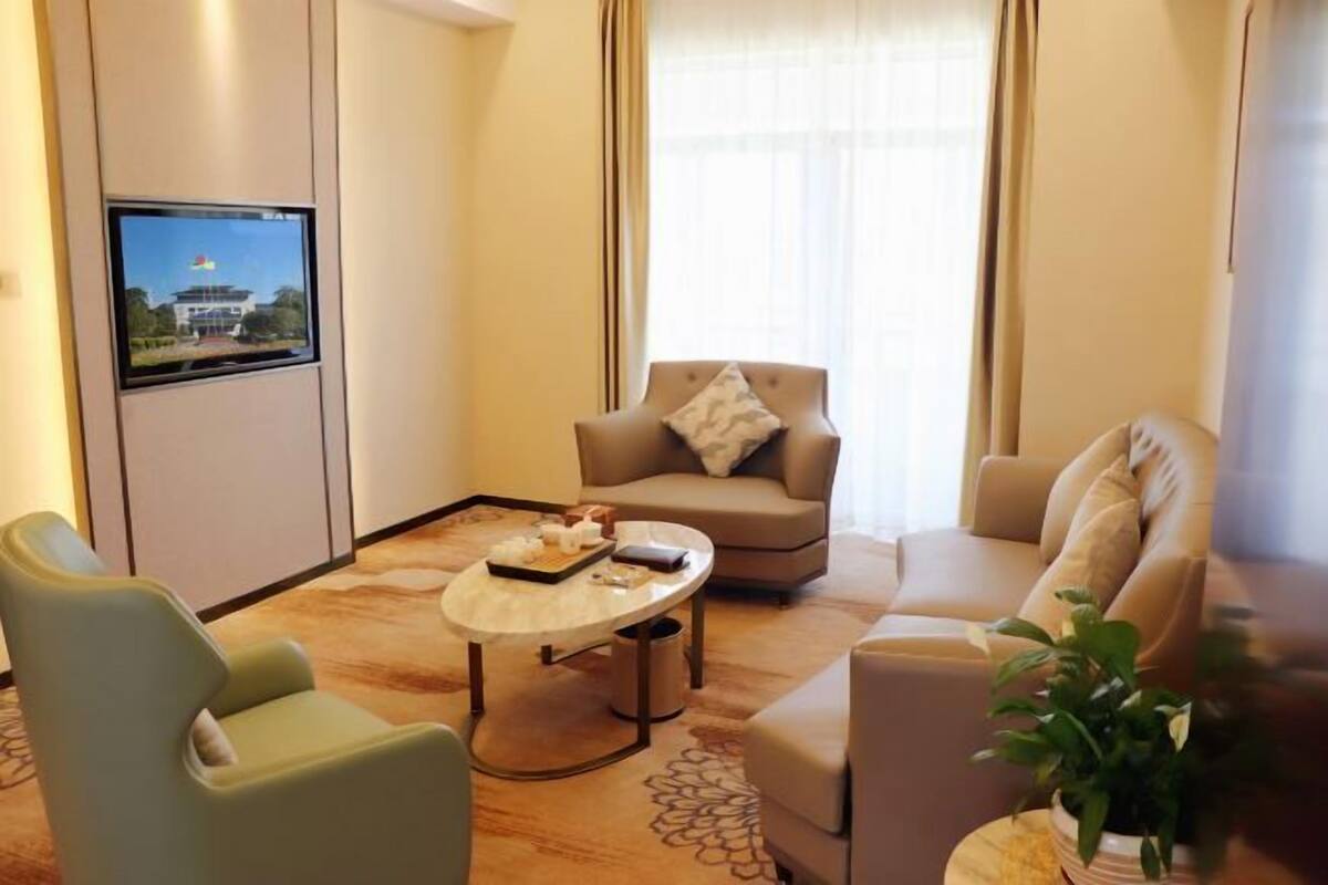 Et moderne hotelværelse med et fladskærms-tv på væggen, en beige sofa, en grøn lænestol og et rundt sofabord med en bakke med snacks.