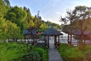 A wooden pathway leading to a gazebo with a thatched roof, surrounded by lush greenery and a body of water. - Shanghang Gutian Hotel (Shanghang)